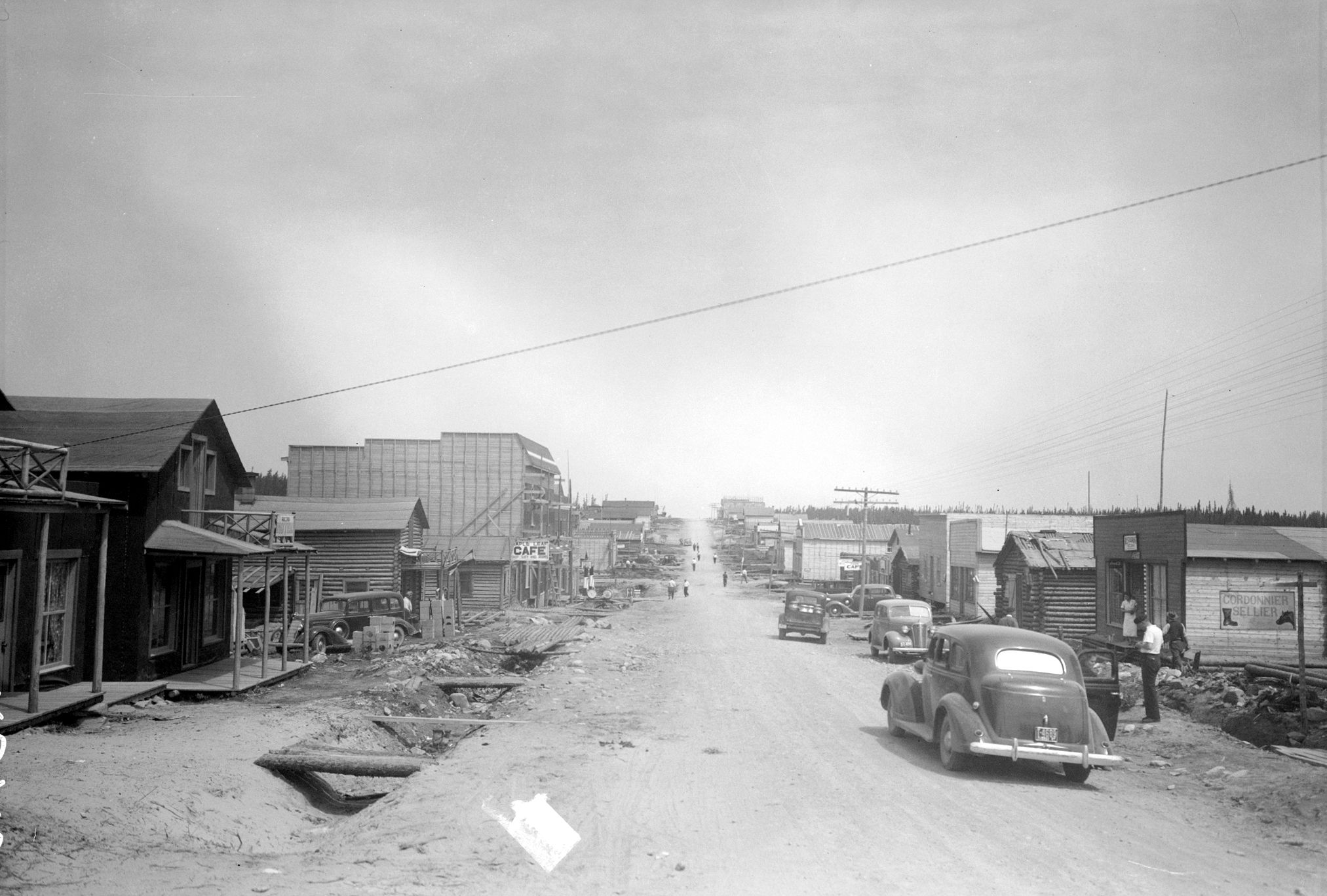 Photographie en noir et blanc d’une route de gravier bordée par des cabanes en bois rond ou de planches. Un fossé sépare les bâtiments et la route sur laquelle plusieurs voitures sont visibles. À droite, une affiche indiquant « Cordonnier sellier » et à gauche une pan-carte « Maple Leaf Café ».