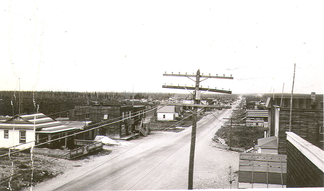 Photographie noir et blanc d’une route de gravier bordé de bâtiments en planches. À l’avant-plan, un poteau supportant des fils. À l’arrière-plan, un champ et une forêt de conifères.  