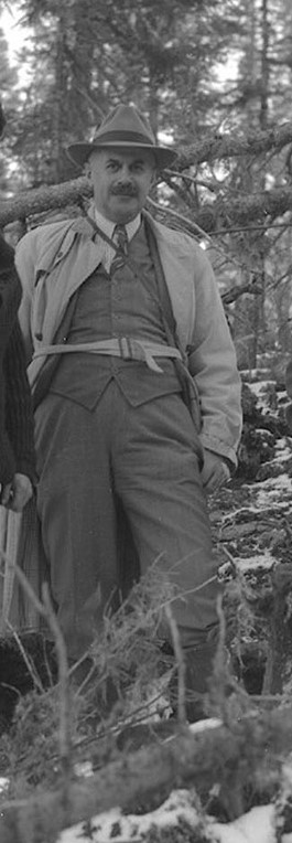 Photographie en noir et blanc d’un homme en habit, avec un chapeau et une moustache en brosse, qui pose dans une forêt. 