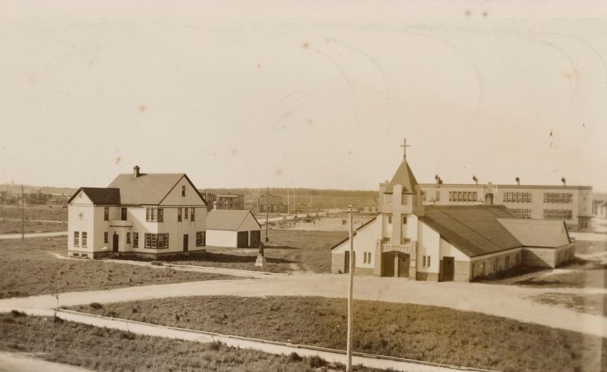 Photographie en sépia d’un presbytère et d’une église. Celle-ci est de faible hauteur et une grosse croix surplombe l’entrée principale. À l’arrière-plan, une école primaire catholique. 