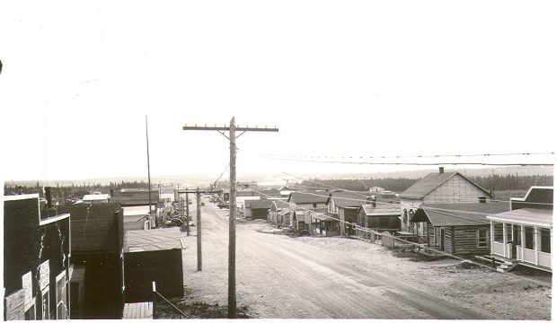 Photographie en noir et blanc d’une route de gravier bordée de bâtiments en bois ou de planches sans doute prise du toit d’un édifice. La forêt est visible à l’arrière-plan.