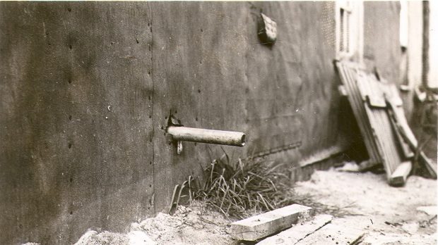 Photographie en noir et blanc d’un tuyau sortant d’un bâtiment recouvert de papier goudronné et se déversant sur des planches de bois. En l’arrière-plan, un tas de bois. 