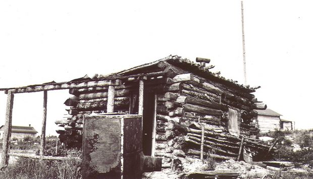 Photographie en noir et blanc d’une cabane en bois rond de construction rudimentaire qui est en train de s’affaisser. En arrière-plan, d’autres bâtiments semblables, mais en meilleur état. 