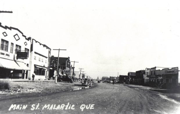 Photographie en noir et blanc d’une large route de gravier bordée d’édifices de bonne qualité. À gauche, les enseignes du magasin « Cleman’s » et d’un hôtel. Plusieurs voitures sont stationnées des deux côtés. À l’arrière-plan, un chevalement minier. 