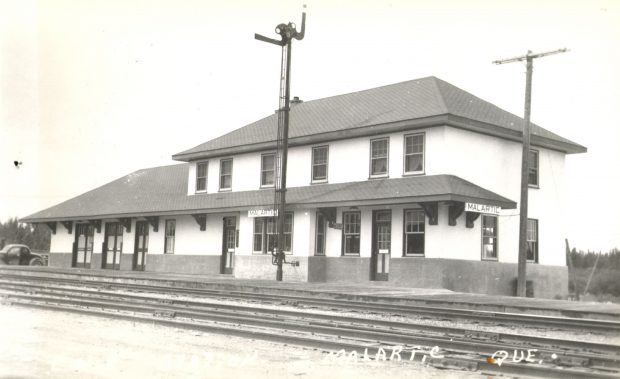 Photographie en noir et blanc d’un bâtiment de deux étages de bonne qualité. À deux endroits, des affiches du nom de la localité « Malartic ». À l’avant-plan, plusieurs voies de chemin de fer. La photo porte l’inscription « Malartic Que ».