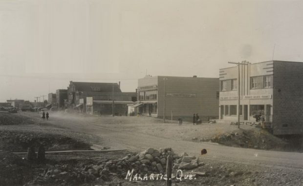 Photographie en noir et blanc d’une route de gravier en construction avec une rangée de bâtiments de construction récente à l’arrière-plan. Deux femmes marchent derrière une voiture qui soulève un nuage de poussière. À l’avant-plan, nous pouvons voir un pont et un petit amas de roches. La photo porte l’inscription « Malartic, Qué ».