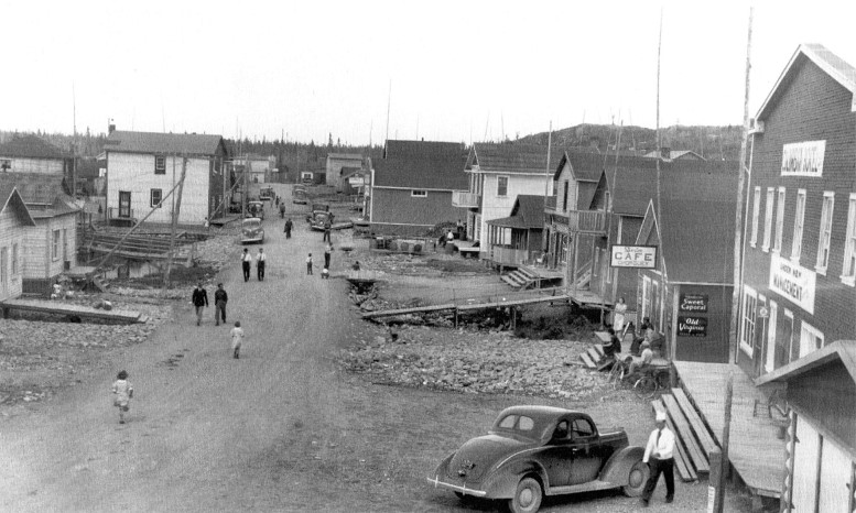 Photographie en noir et blanc d’une étroite route de gravier bordée de bâtiments rudimen-taires. Plusieurs voitures sont stationnées et une dizaine de personnes circulent sur la voie publique. De longs trottoirs de bois permettent d’accéder à certaines résidences.