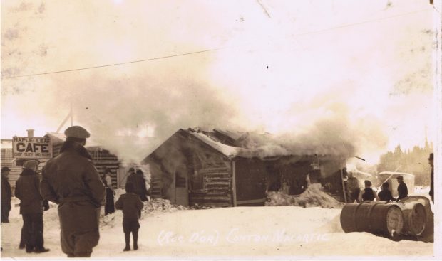 Photographie en sépia d’une cabane en bois rond alors qu’elle est incendiée. Un large nuage de fumée occupe la moitié supérieure de la photo. Une douzaine de curieux assiste à la scène. À gauche, l’enseigne « Maple Leaf CAFE Chop Suey and Rooms ».