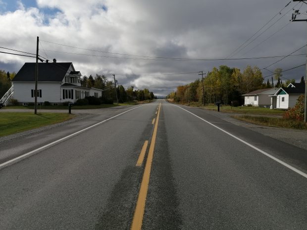 Photographie en couleur d’une route en asphalte avec quelques résidences sur les côtés. À gauche, une maison blanche de deux étages et deux résidences à droite. Le ciel est nuageux. 