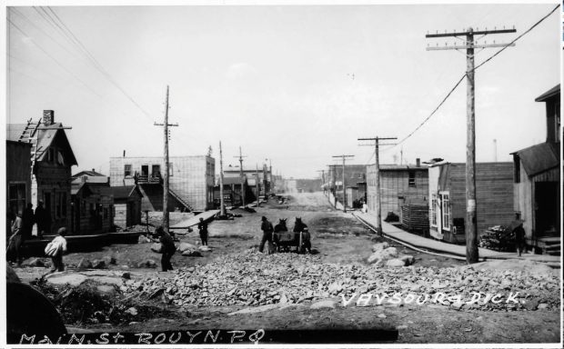 Photographie en noir et blanc d’une rue bordée de bâtiments rudimentaires et de poteaux de lignes de distribution d’électricité. Six hommes, ainsi que deux chevaux, travaillent au pavage. Des trottoirs de bois longent la rue. 