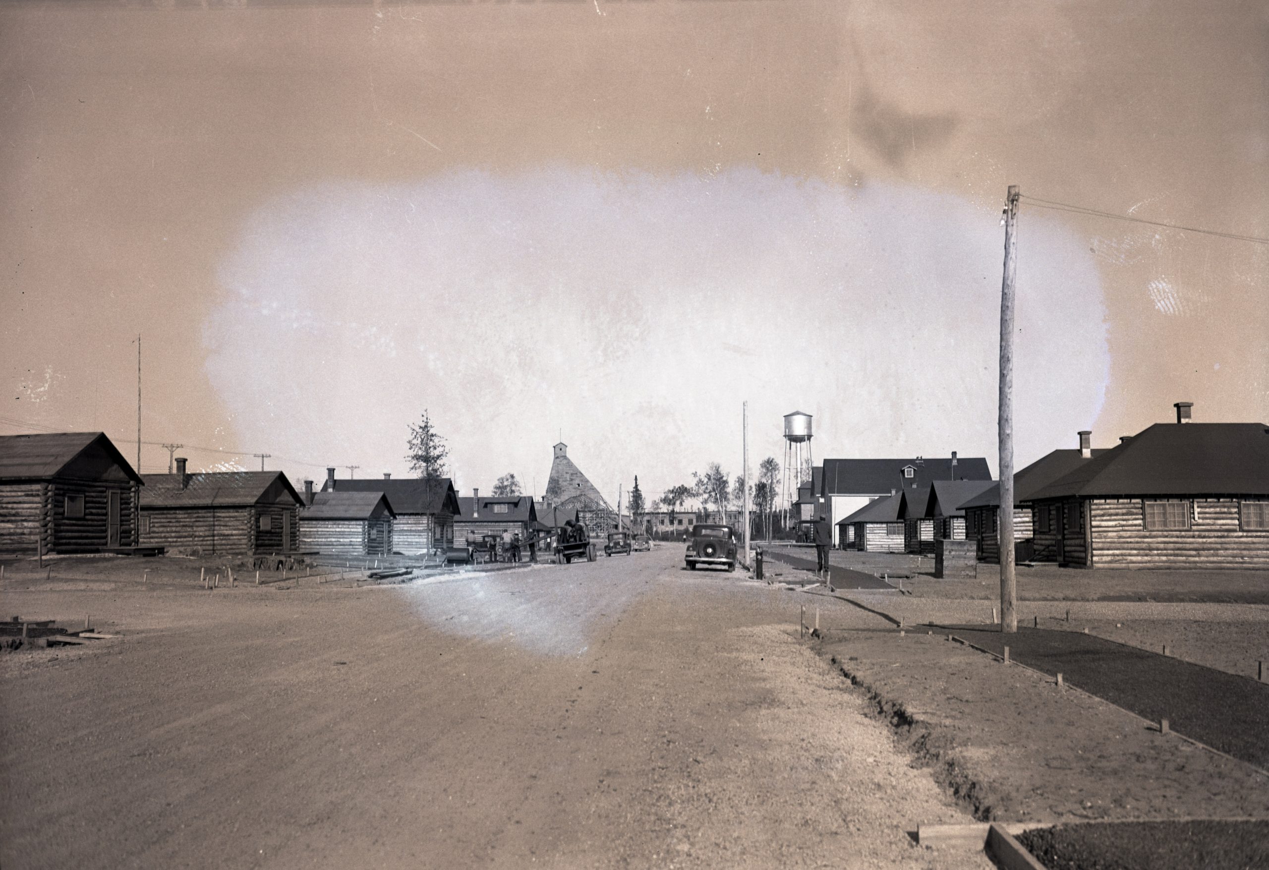 Photographie en noir et blanc d’une rue bordée de cabanes en bois rond. Des hommes travaillent à la construction des trottoirs. En arrière-plan, un château d’eau et un chevalement minier. 