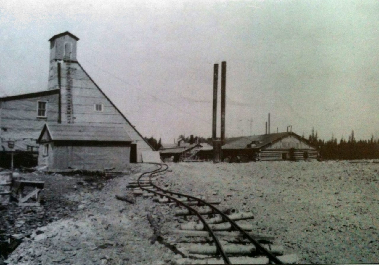 Photographie en noir et blanc d’un chevalement minier et d’un bâtiment rudimentaire surmonté de deux cheminées. À l'avant-plan, un chemin de fer sinueux.
