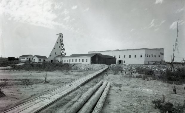 Photographie en noir et blanc des infrastructures de la mine Canadian Malartic, dont un chevalement de planches. À l’avant-plan, des billots de bois et un sentier de planches permettant d’accéder aux bâtiments. 