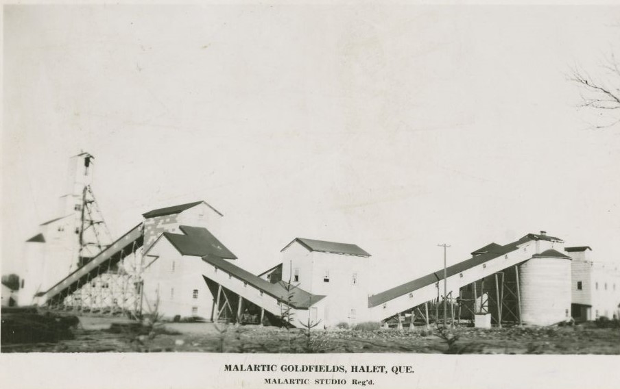 Photographie en noir et blanc de bâtiments industriels, dont un chevalement de mine et plusieurs convoyeurs. En bas, l’inscription « Malartic Goldfields, Halet, Que. Malartic Strudio Reg’d ».