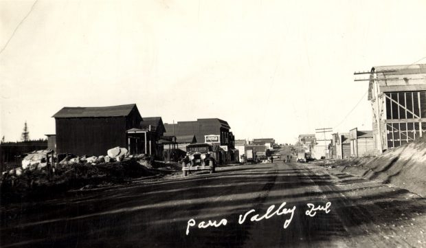 Photographie en noir et blanc d’une route bordée de bâtiments de planches sur laquelle plusieurs piétons circulent. À l’avant-plan, une voiture de taxi. En bas, il est inscrit en blanc « Paris Valley Que ».