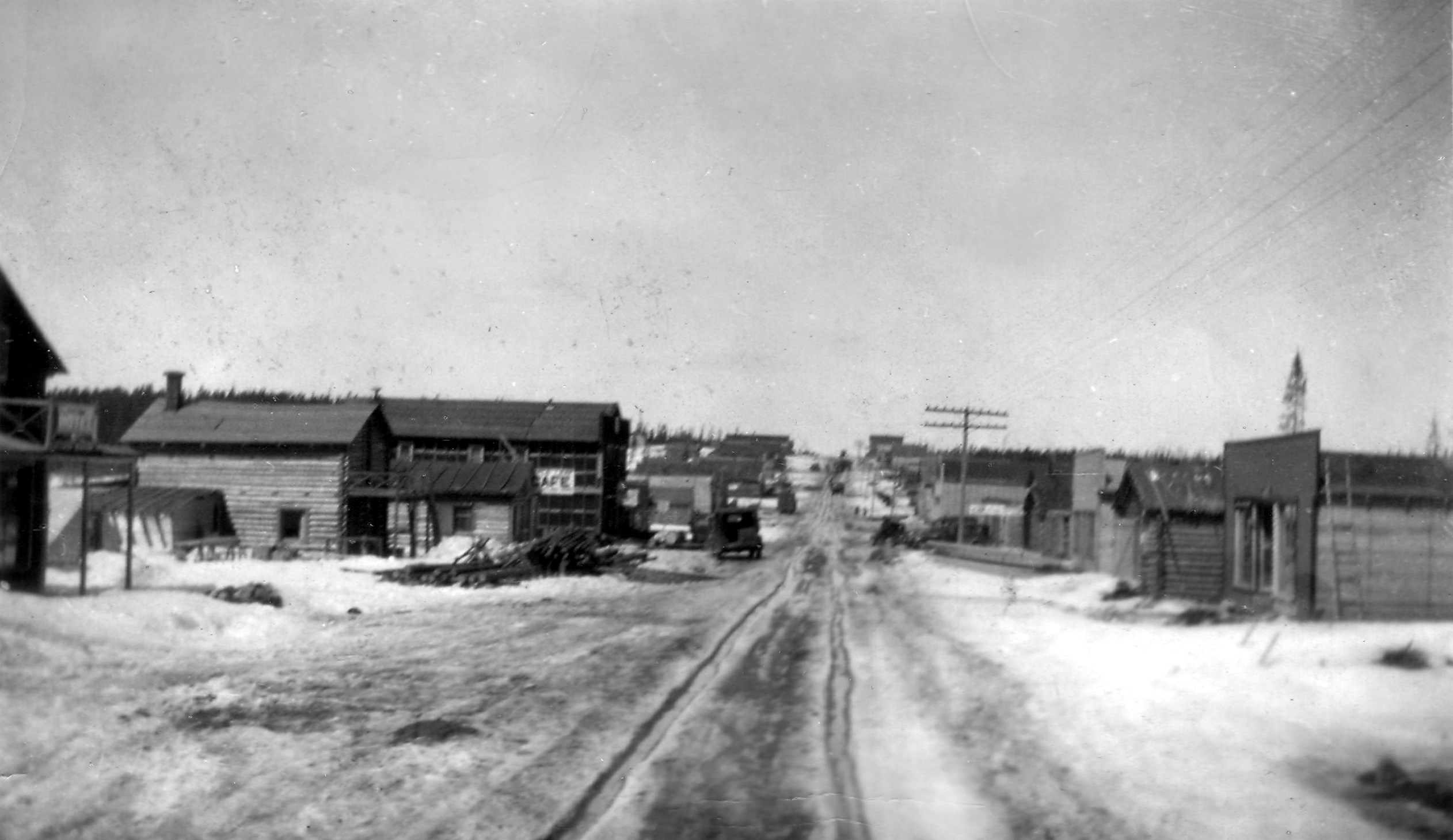 Photographie en noir et blanc d’une route en hiver bordée de bâtiments de planches ou en bois rond. Vu la circulation dans la neige, des roulières sont visibles au centre. 