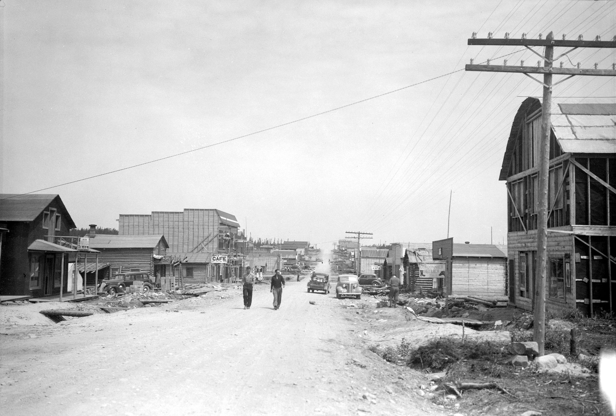 Photographie en noir et blanc d’une route de gravier bordée de bâtiments de planches ou en bois rond. Deux hommes marchent vers la caméra. À l’avant-plan, à droite, un poteau téléphonique. 