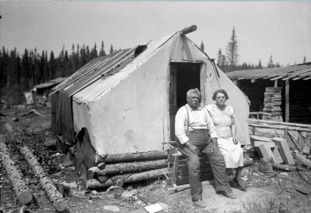 Photographie en noir et blanc d’un couple de personnes âgées devant une habitation rudimentaire, soit une tente en bois et en textile. Des cabanes en bois rond sont visibles à droite et à l’arrière-plan.