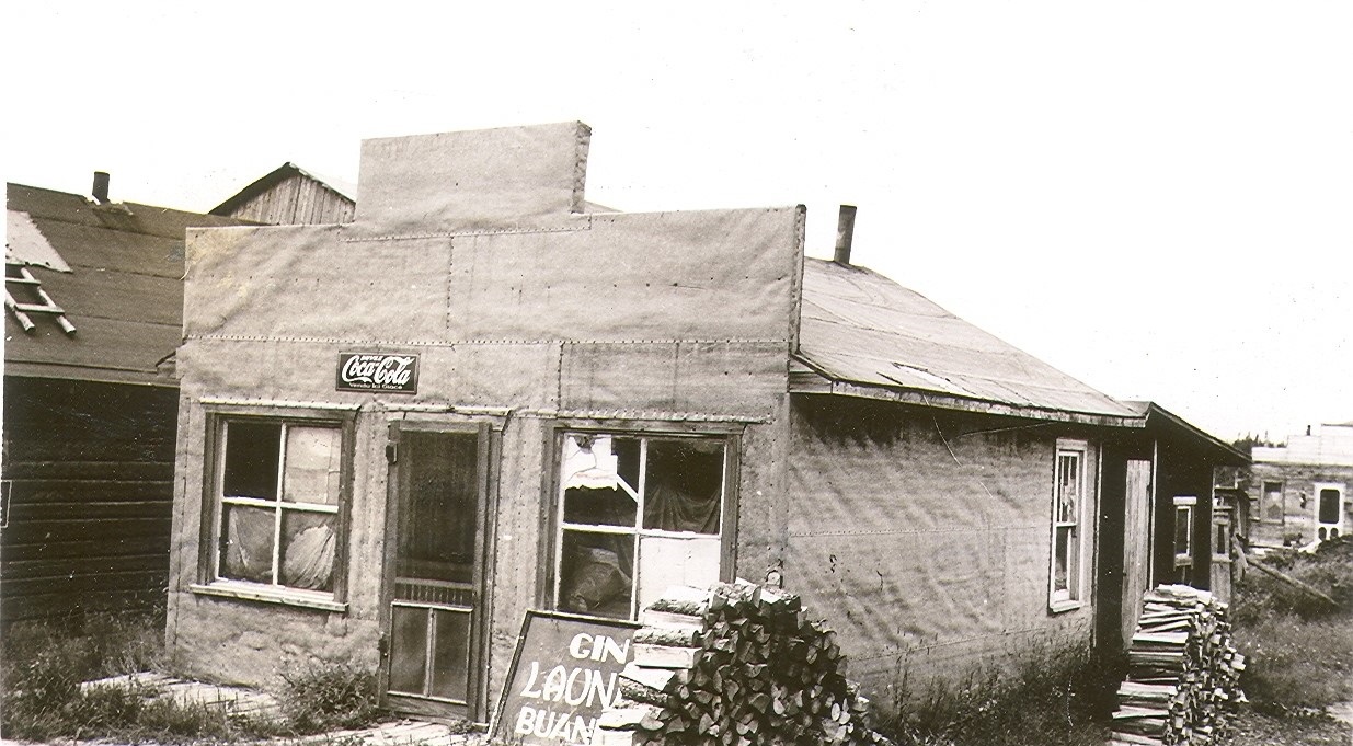Photographie en noir et blanc d’une habitation en papier goudronné avec une façade de style Boomtown. Un enseigne est déposé au sol : « Gin LAUNDRY buanderie ». 
