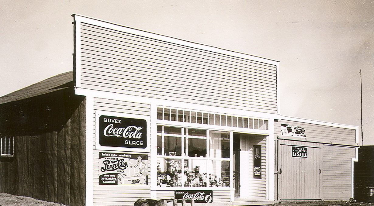 Photographie en noir et blanc d’une habitation de planches avec façade de style Boom-town sur laquelle plusieurs publicités « Coca Cola » sont affichées. Plusieurs produits sont présentés en vitrine. 