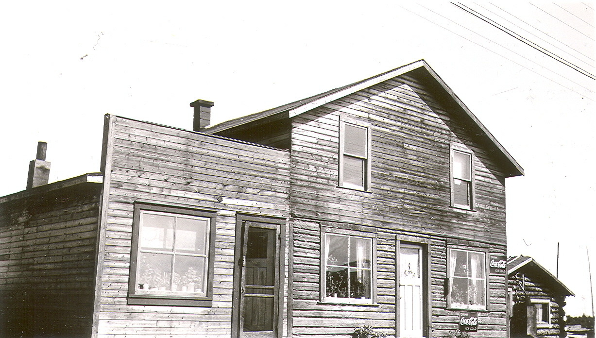 Photographie en noir et blanc d’un immeuble de planches contenant deux logements. Deux publicités de « Coca-Cola » sont affichées sur la résidence. À droite, une cabane en bois rond. 