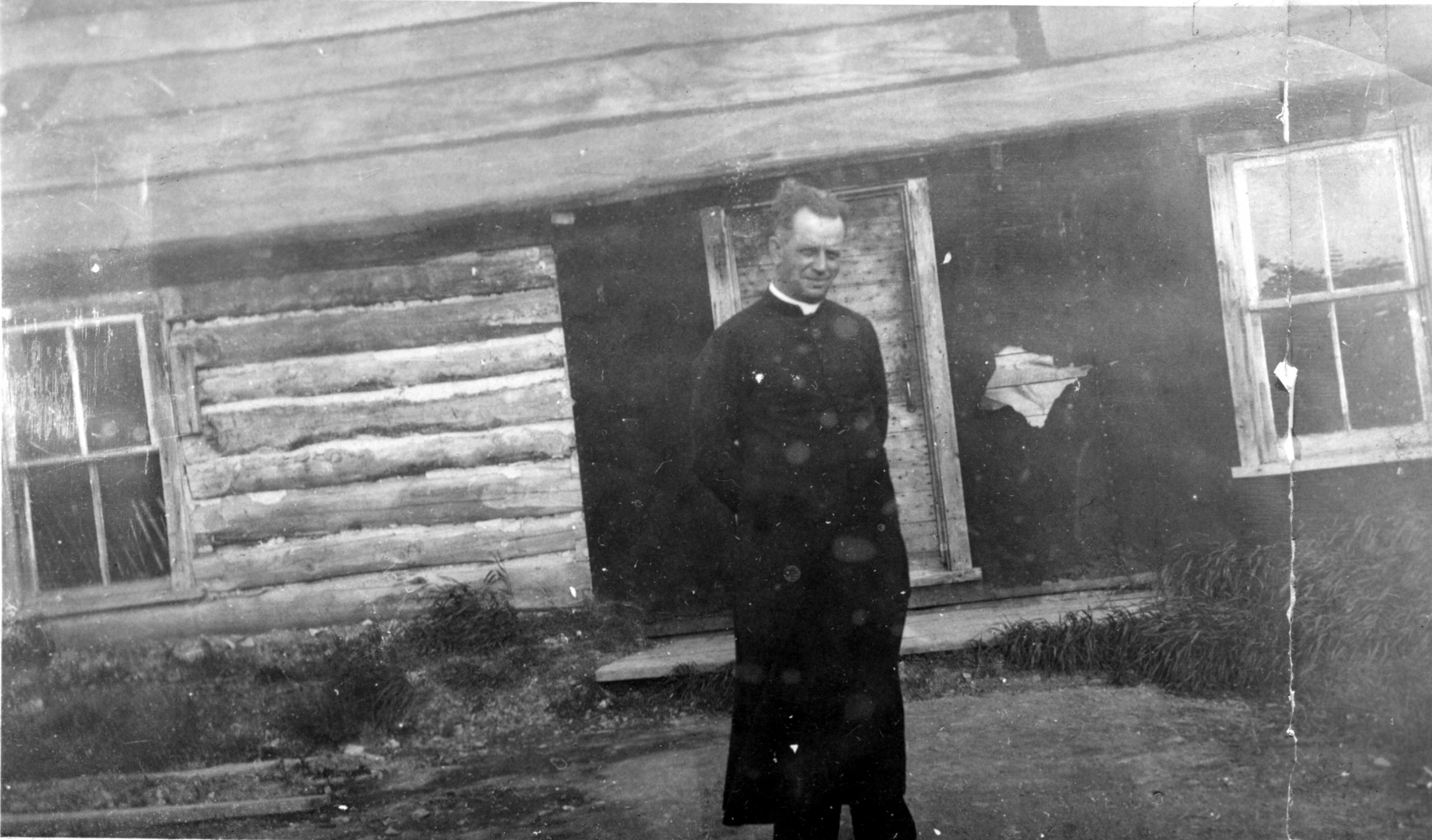 Photographie en noir et blanc d’un homme en soutane qui pose devant une cabane en bois rond recouverte partiellement de papier goudron. 