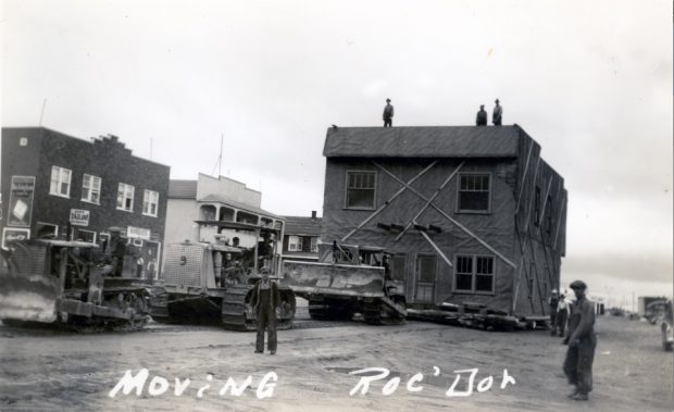 Photographie en noir et blanc d’un bâtiment de deux étages déplacé par trois tracteurs. Plusieurs hommes sont sur le toit. Plusieurs curieux assistent à la scène. À l’arrière-plan, plusieurs immeubles. 
