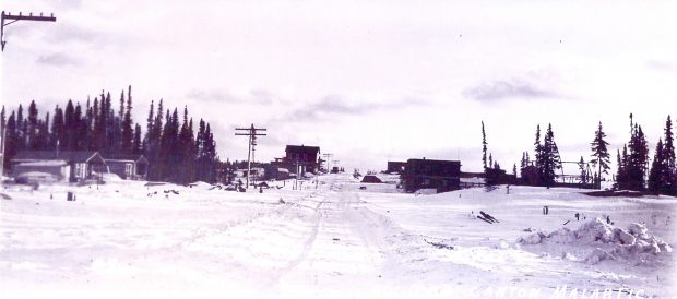 Photographie noir et blanc d’un hameau enneigé avec une route au centre. Des bâtiments en bois rond et en planche et des poteaux de téléphone sont visibles. 