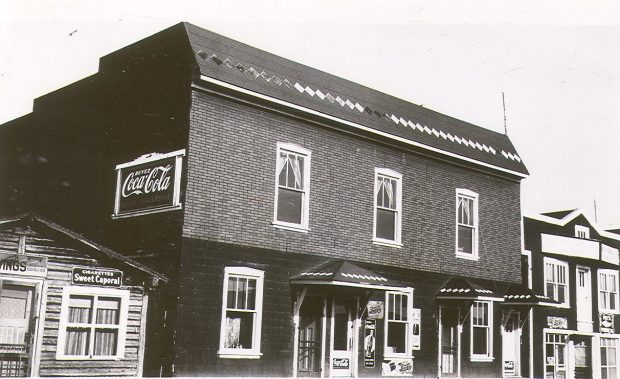 Photographie en noir et blanc d’un bâtiment de deux étages recouvert de papier goudronné. La devanture a six fenêtres et quatre portes. Sur le côté, une publicité « Buvez Coca-Cola ». À droite, l’hôtel Frontenac et une cabane en bois rond à gauche. 
