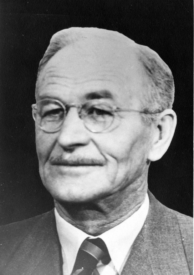 Photographie en noir et blanc d’un homme en habit-cravate qui porte des lunettes et une mince moustache. 