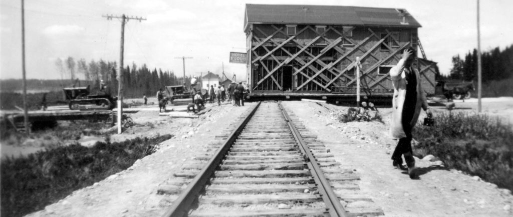 Photographie en noir et blanc d’un bâtiment de deux étages qui traverse un chemin de fer. L’enseigne « Hotel St-Louis » est visible. Deux tracteurs tirent l’édifice et une dizaine de curieux assistent à la scène. 