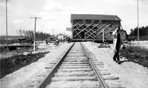 Photographie en noir et blanc d’un bâtiment de deux étages qui traverse un chemin de fer. L’enseigne « Hotel St-Louis » est visible. Deux tracteurs tirent l’édifice et une dizaine de curieux assistent à la scène. 