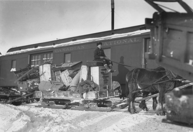 Photographie noir et blanc d’une charrette, remplie à ras bord de meubles et de boites, qui est tirée par deux chevaux conduits par un homme assis sur les biens. Vu la neige, la charrette est sur des skis et l’homme est habillé chaudement. À l’arrière-plan, un wagon portant l’inscription « Canadian National ». 