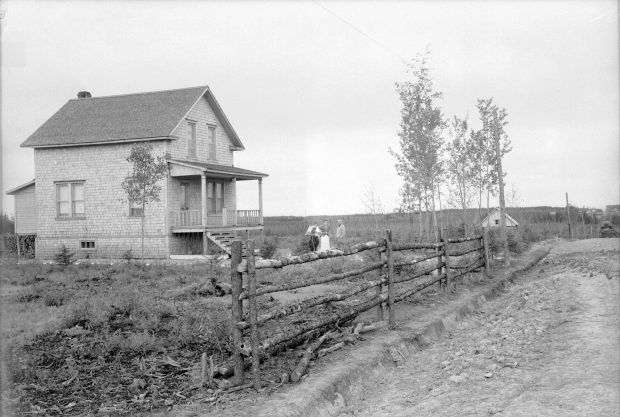 Photographie en noir et blanc d’une maison de deux étages avec trois personnes qui po-sent devant. À l’avant-plan, une clôture. À l’arrière-plan, une petite agglomération de maisons. 