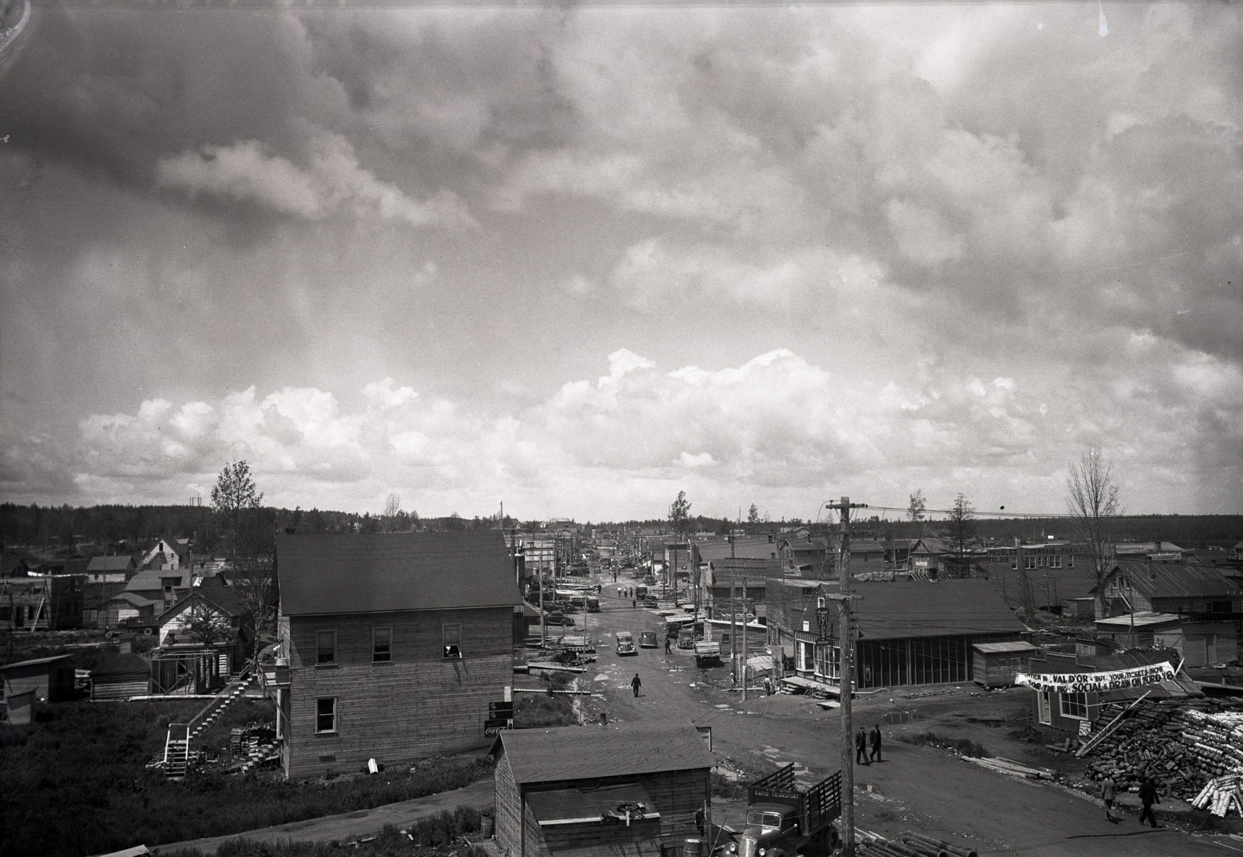 Photographie en noir et blanc d’une rue bordée des deux côtés de bâtiments rudimentaires. Des voitures et des piétons circulent au centre de la route non pavée qui est parsemée de trous d’eau. 