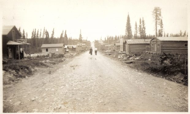 Photographie en sépia d’une route de gravier bordée de cabanes en bois rond. Au centre, deux hommes marchent vers une colline et plusieurs conifères sont visibles à l’arrière-plan.