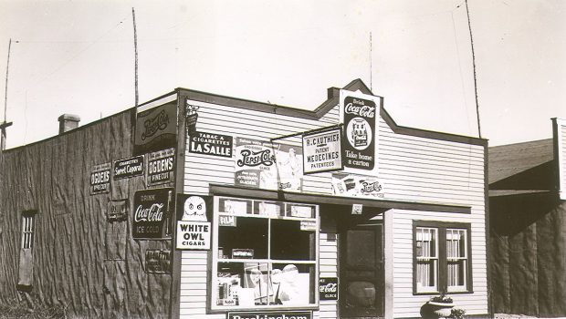 Photographie en noir et blanc d’un bâtiment dont la façade est recouverte de planches et le côté de papier goudronné. Une douzaine de publicités sont affichées, notamment pour Coca-Cola et pour différentes marques de cigarettes (Sweet Caporal et Lasalle). 