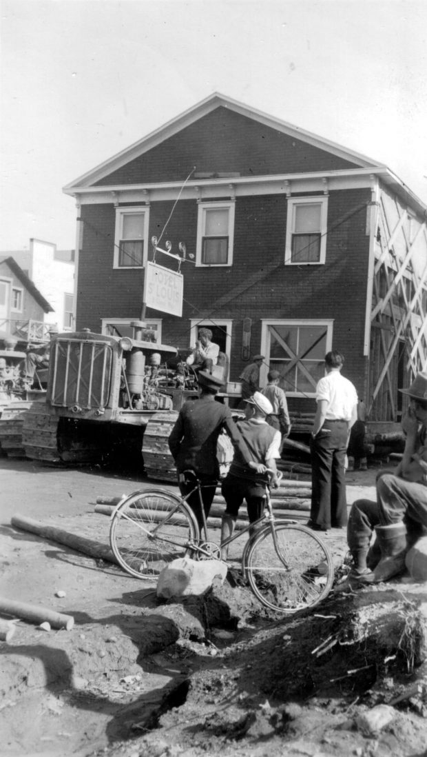 Photographie  en noir et blanc d’un bâtiment de deux étages déplacéer par un tracteur. Plusieurs curieux, dont un avec sa bicyclette, assistent à la scène. 