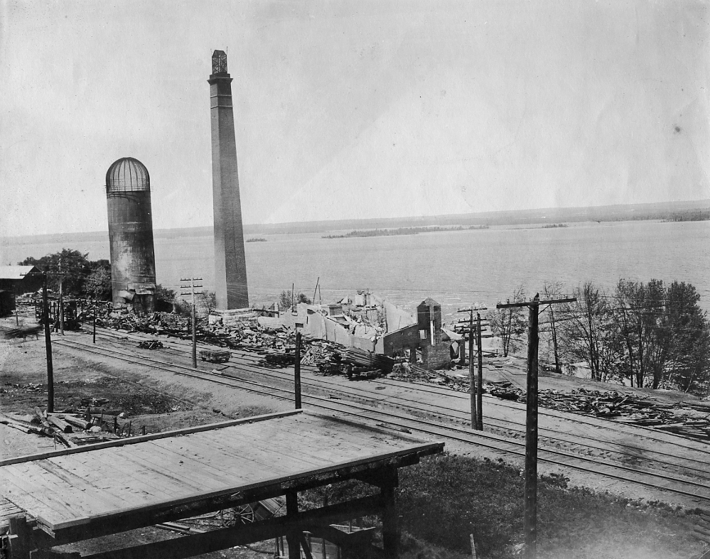 Two chimneys stand among the ruins of burned out buildings next to a river.