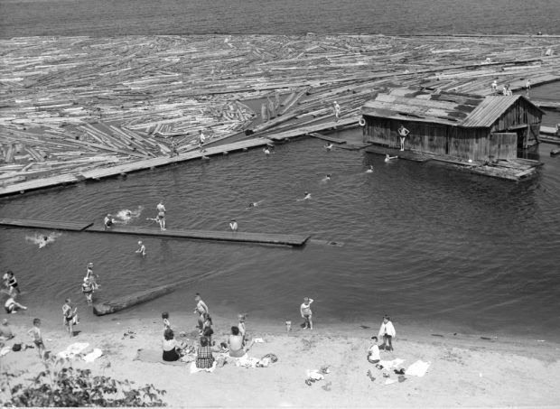 Des femmes assises sur une plage sablonneuse surveillent des enfants qui nagent jusqu’à un hangar à bateaux, non loin de la rive.