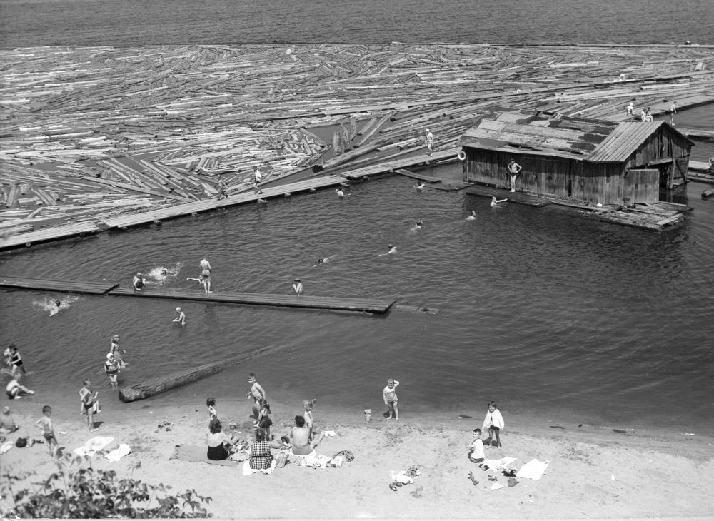 Des femmes assises sur une plage sablonneuse surveillent des enfants qui nagent jusqu’à un hangar à bateaux, non loin de la rive.