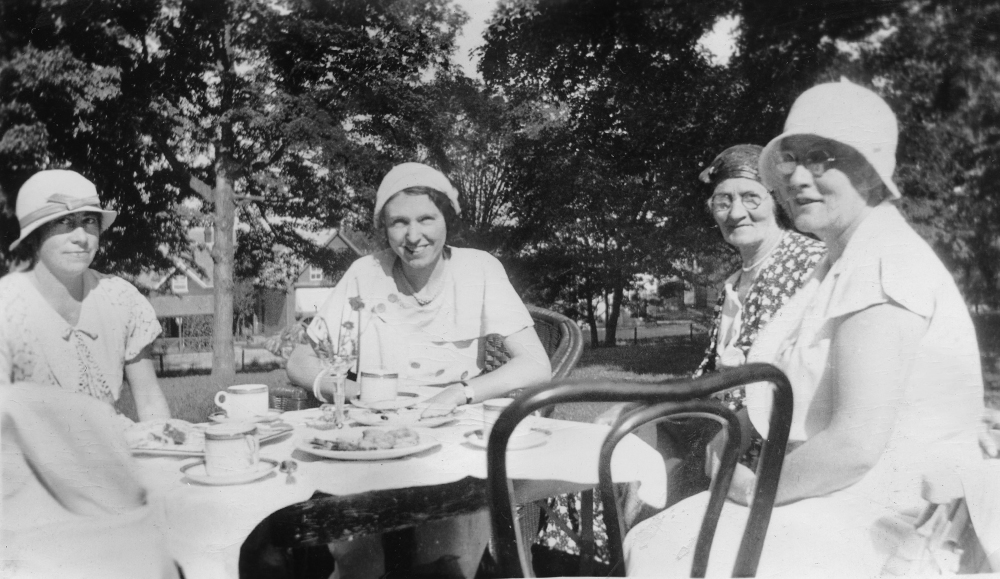 Quatre femmes coiffées de chapeaux prennent le thé autour d’une table et sourient au photographe.