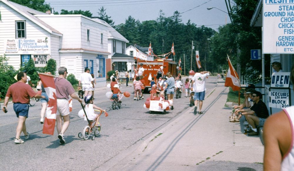 Un camion de pompier décoré mène un défilé d’enfants à vélo et d’adultes qui brandissent des drapeaux canadiens.
