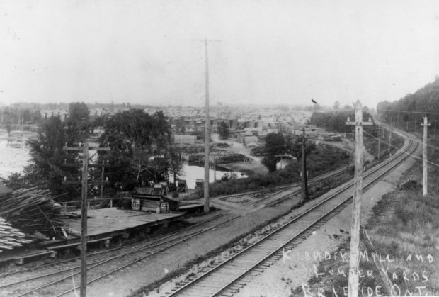 Une photo en noir et blanc montre des piles de bois d’œuvre qui s’étendent le long d’une voie ferrée.