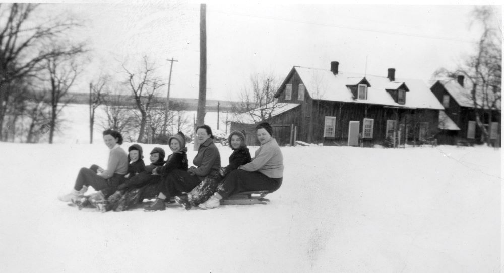 Des femmes et enfants sont assis sur un long traîneau au sommet d’une colline et sourient à la caméra.