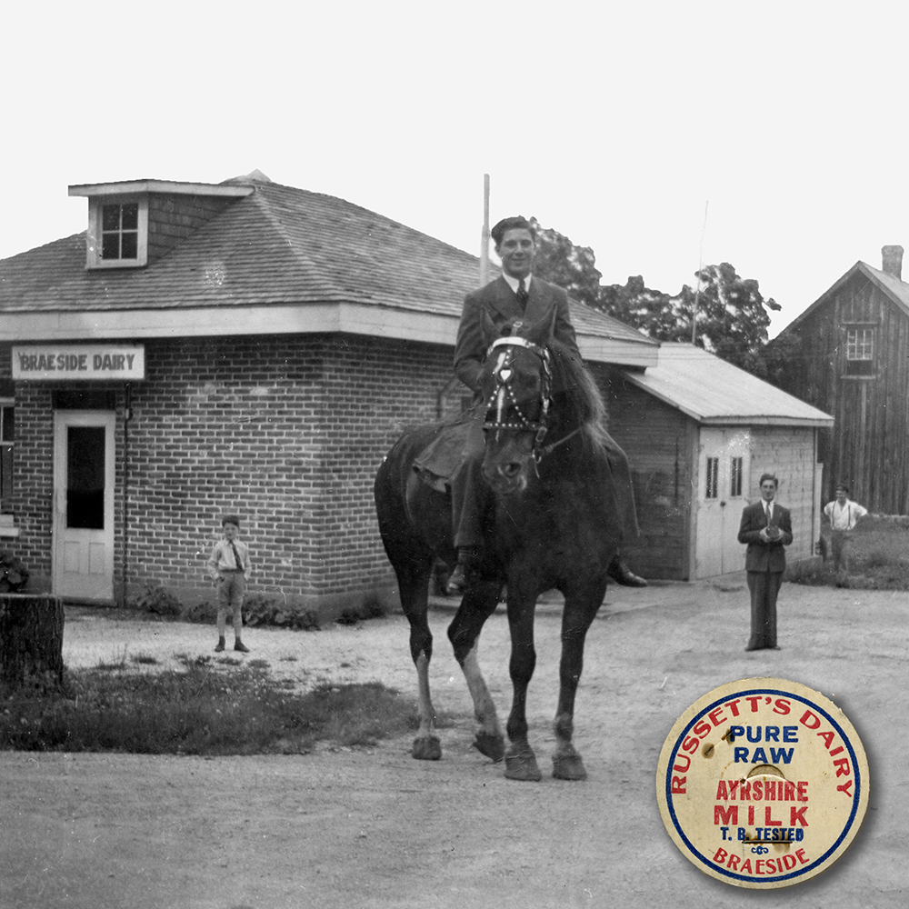 Un jeune homme monte un cheval devant une petite bâtisse dont l’enseigne affiche « Laiterie de Braeside ». Un jeune garçon et deux autres hommes se tiennent à l’écart et regardent vers le photographe. Un médaillon superposé apparaît dans le coin inférieur droit de la photo, montrant le bouchon d’une bouteille de lait Russett.