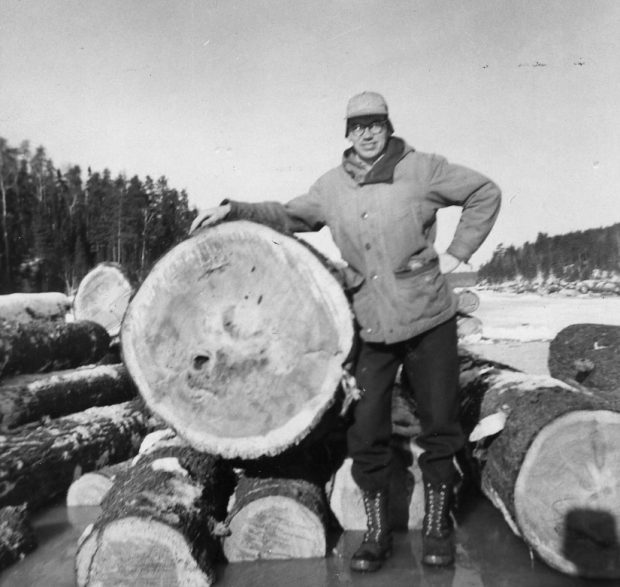 Un forestier se tient debout sur la glace avec une main plantée sur la hanche et l’autre posée sur une énorme bille de bois.