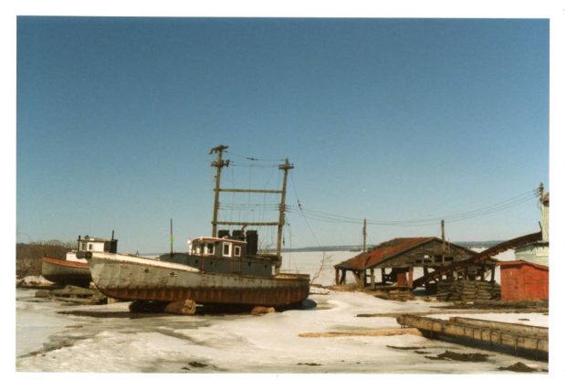 Deux vieux remorqueurs reposent sur des billes de bois à côté d’un hangar de bois délabré au bord de l’eau en hiver.