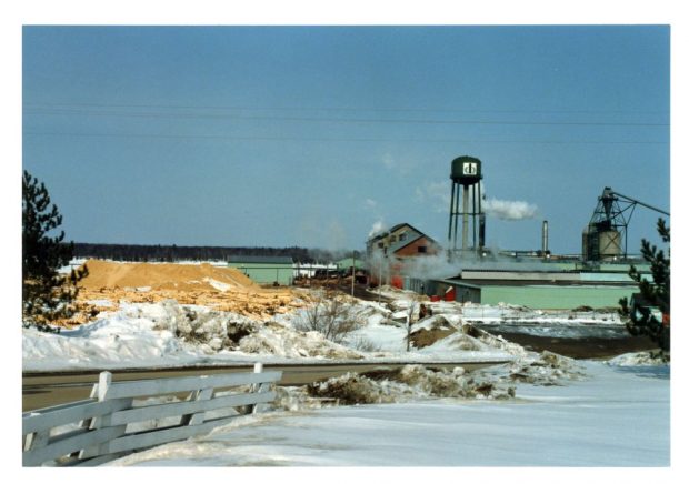 Une scierie faite de briques est entourée de piles de bran de scie l’hiver, avec un château d’eau et un incinérateur à déchets en arrière-plan.