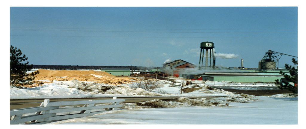 Une scierie faite de briques est entourée de piles de bran de scie l’hiver, avec un château d’eau et un incinérateur à déchets en arrière-plan.